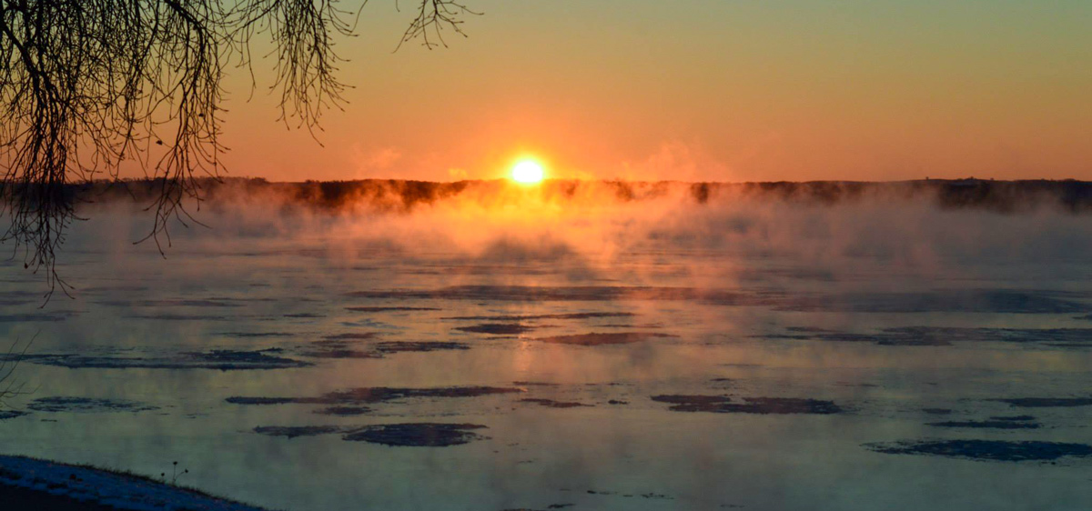 The sun makes an orange ball over a dark river flowing with ice